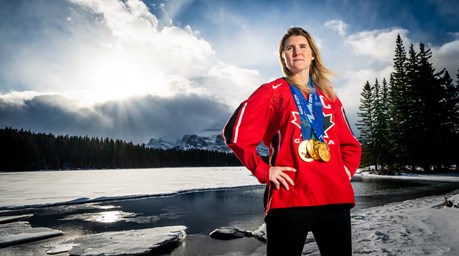 Hayley Wickenheiser in front of mountains wearing a team Canada jersey and 4 Olympic gold medals.